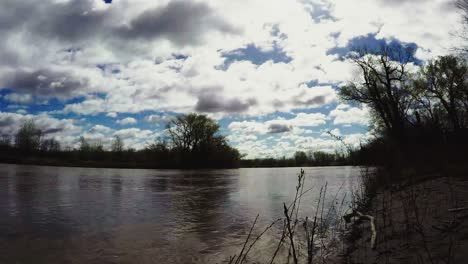 Timelapse-De-La-Orilla-Del-Río-De-Principios-De-La-Primavera-De-Dos-Niveles-De-Nubes-En-Un-Día-Soleado-Con-árboles-Y-Ramas-En-La-Orilla-Del-Río