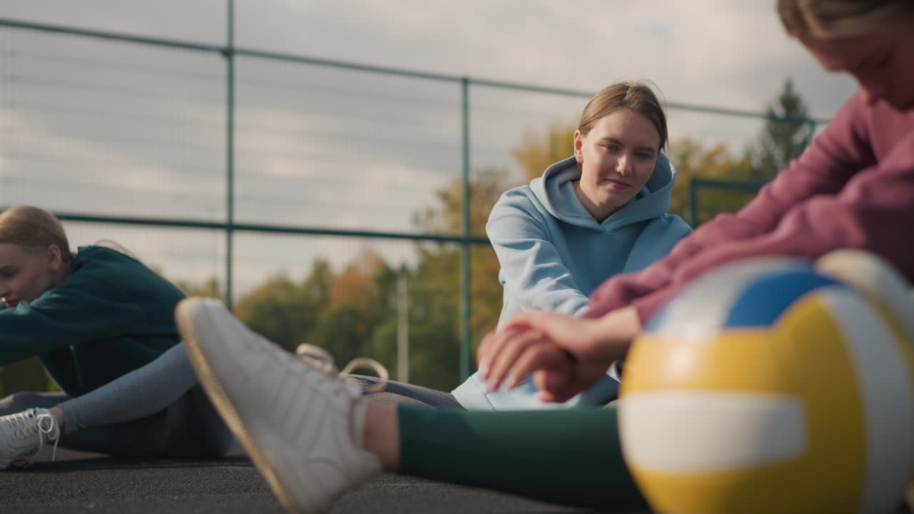 3명의 여성들이 배구 공을 들고 스트레칭을 하고 있는 장면, 다른 여성들이 야외 스포츠 경기장에서 배경에서 운동을 하고 있는 장면, 피트니스 훈련과 팀워크를 보여주는 장면.