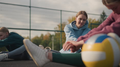 primer plano de tres damas estirándose con una pelota de voleibol cerca, mientras que otras trabajan en el fondo en una arena deportiva al aire libre, mostrando entrenamiento de fitness y trabajo en equipo