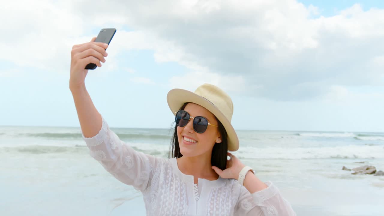 vista lateral de una joven mujer caucásica tomando una selfie con un teléfono móvil en la playa 4k