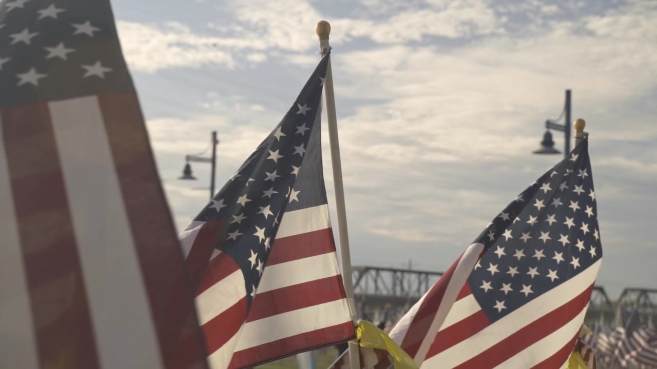 Tre bandiere americane sventolano in primo piano in un parco commemorativo. Questa scena patriottica celebra il Giorno dell'Indipendenza con una profondità di campo ridotta, mettendo in risalto il tessuto contro il cielo luminoso.