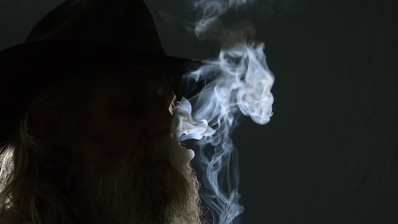 Bearded man in Stetson hat smokes cigarette, backlit in dark room