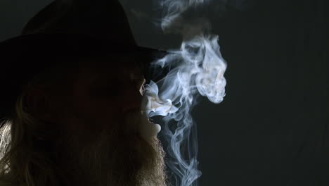 Bearded man in Stetson hat smokes cigarette, backlit in dark room