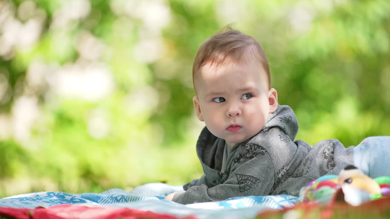 Serious little baby boy lies on the green grass. Cute kid looks into camera and then behind him. Blurred nature backdrop.