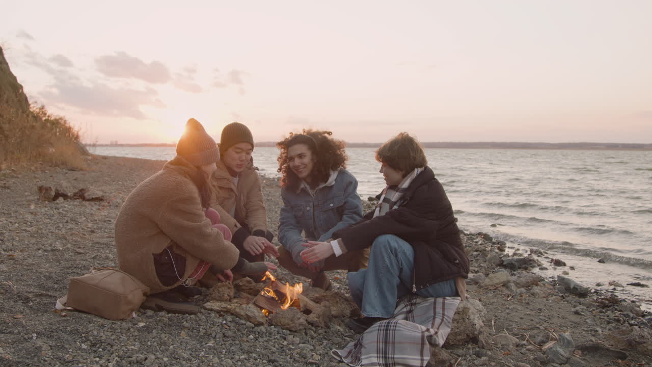 Group Of Teenage Friends Talking And Warming Their Hands At The Bonfire Next To The Seashore