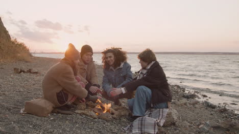 group of teenage friends talking and warming their hands at the bonfire next to the seashore