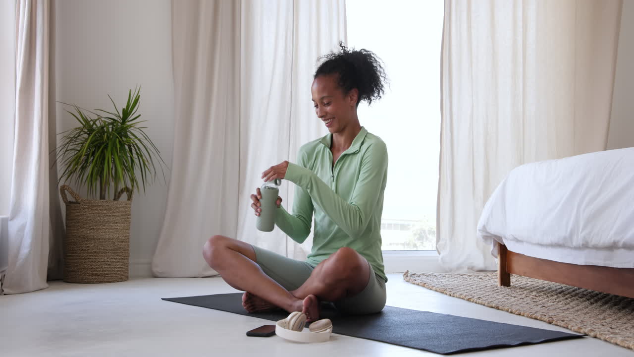 Woman in green activewear drinking water on yoga mat at home, relaxing after workout