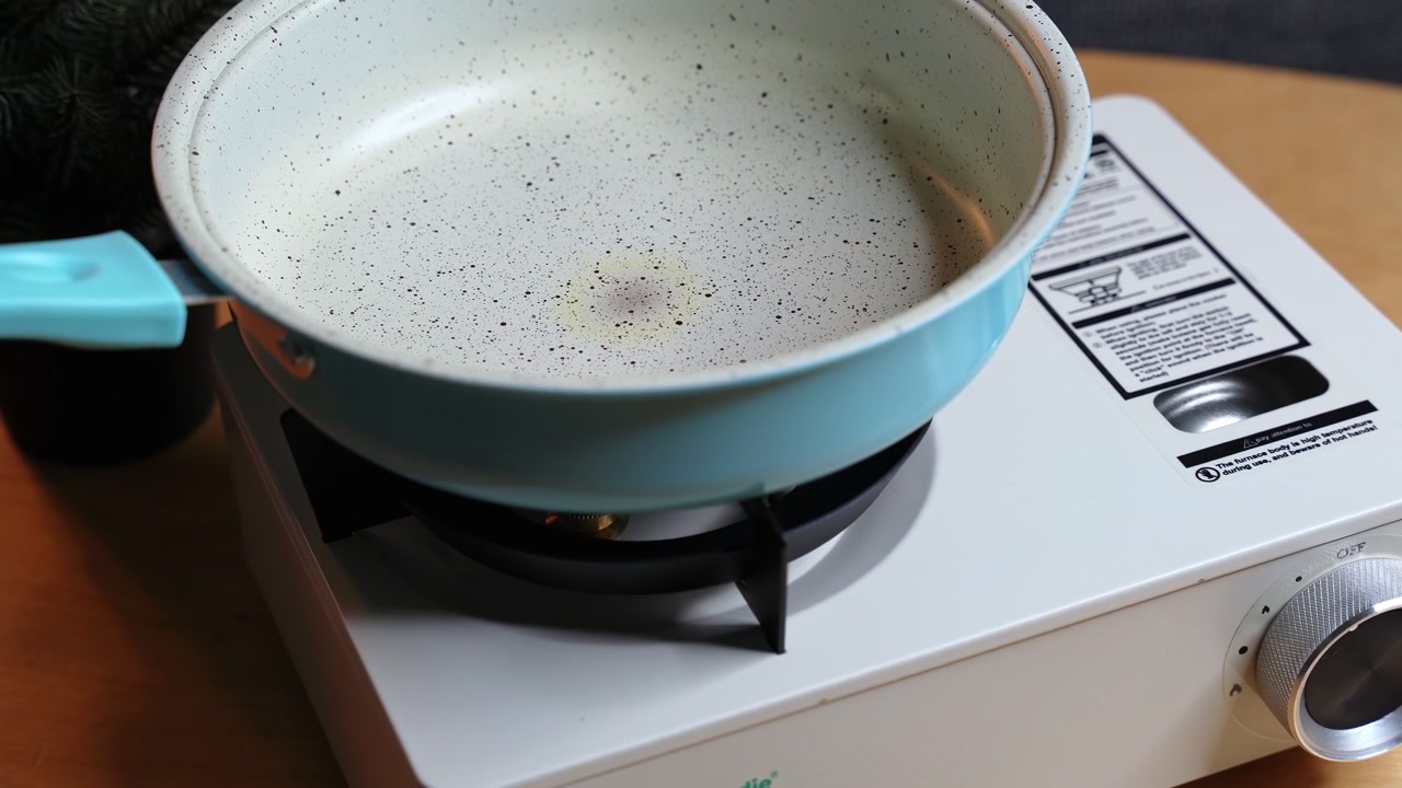Close-up of a blue non-stick frying pan heating on a white portable gas stove top. The skillet rests on a tabletop burner in an indoor kitchen setting, ready for cooking prep.