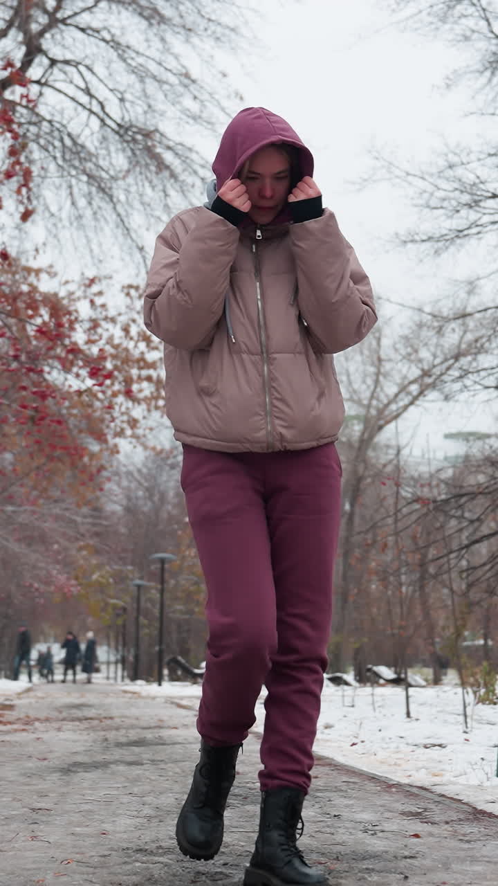 joven vestida con ropa cálida de invierno camina por el camino nevado del parque, sosteniendo la capucha para el calor, árboles desnudos con bayas rojas enmarcan el camino helado, mientras que las figuras distantes borrosas