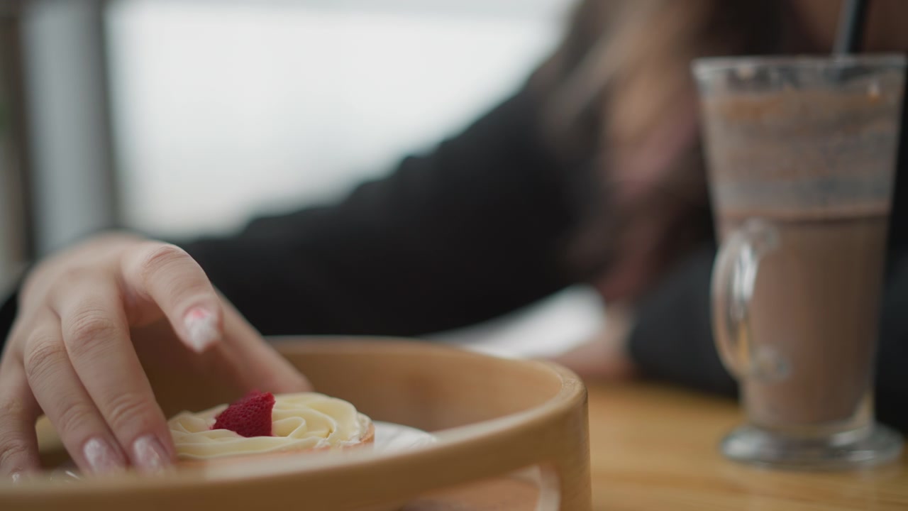 Visible hand lifts strawberry cream pastry from wooden tray beside tall drink in cozy cafe interior, with shallow focus, warm light, relaxed brunch atmosphere, and gentle serving moment at tabletop