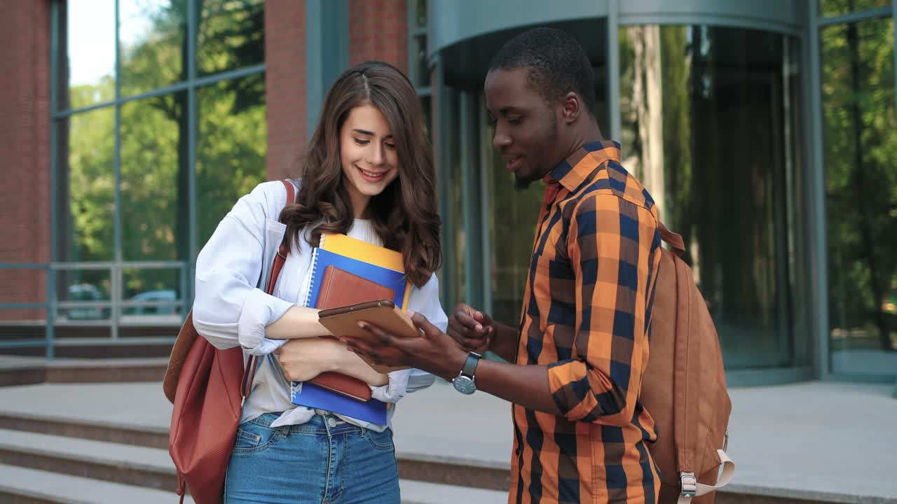 mujer caucásica y hombre afroamericano hablando y viendo algo en la tableta en la calle cerca de la universidad