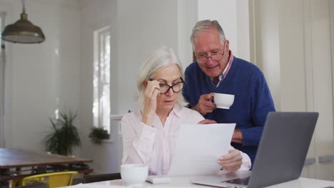 pareja de ancianos con taza de café usando computadora portátil y comprobando las finanzas en casa