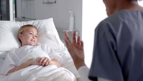 diverse female doctor and girl patient lying on bed talking in hospital, slow motion