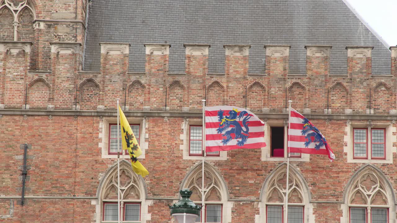 Three colorful flags wave in wind on medieval brick facade, overcast daylight, static camera