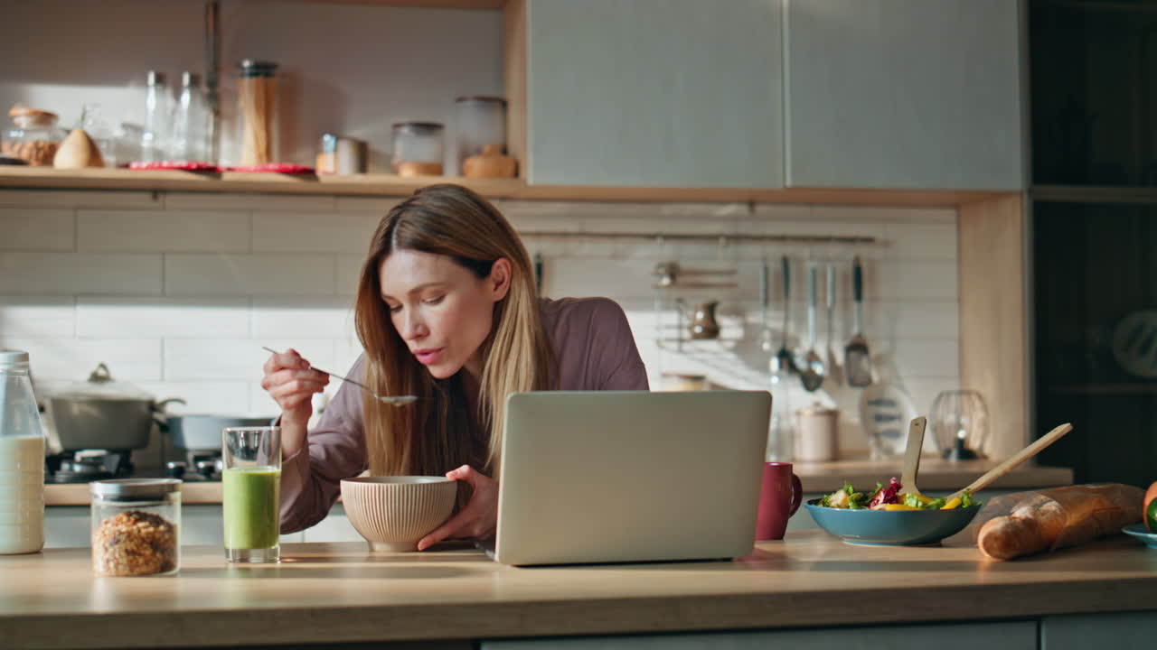 Breakfasting woman looking laptop screen on kitchen. Relaxed girl eating food