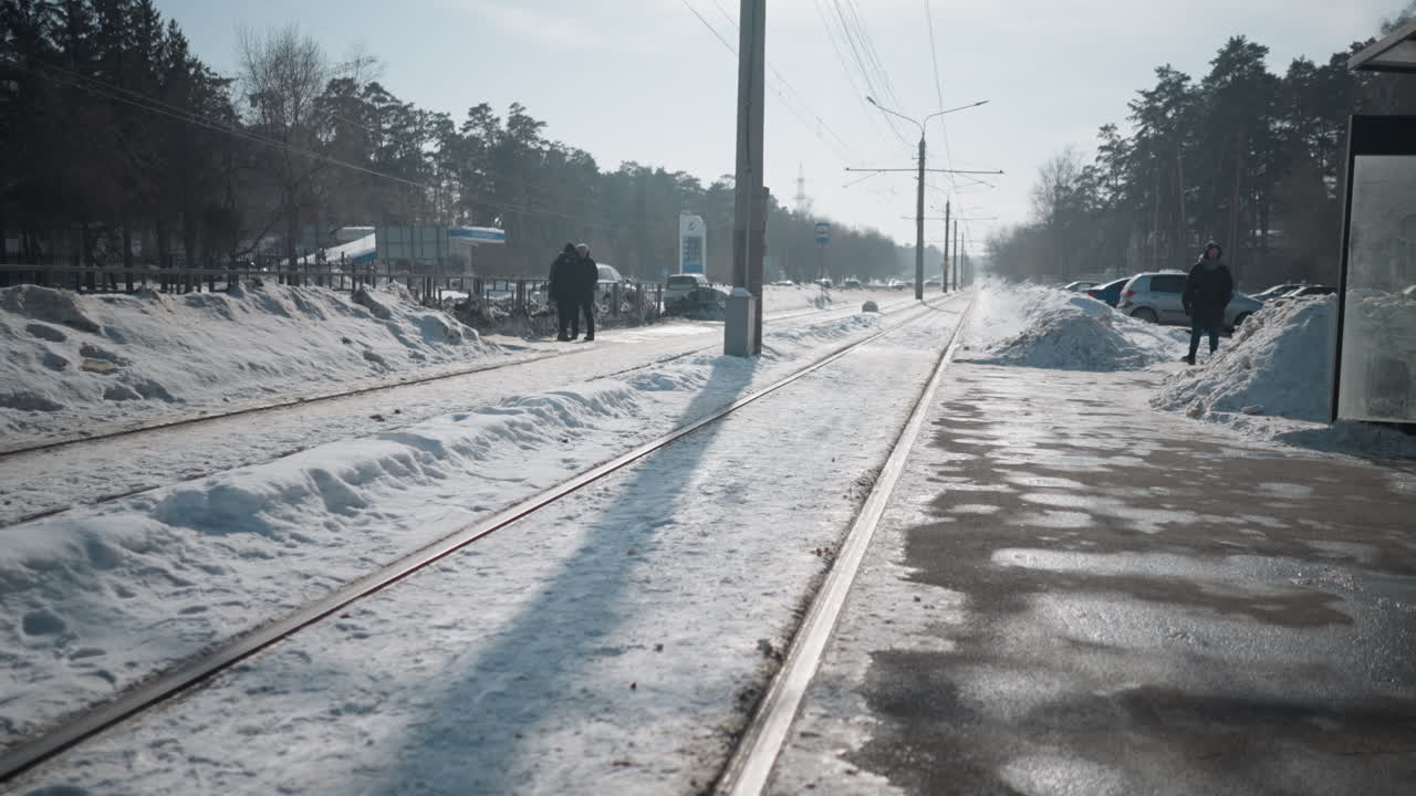 Zoom view pedestrians walking along path past train stop beside snowy tram tracks, cars move through traffic lane while other cars parked near shelter, long morning shadows