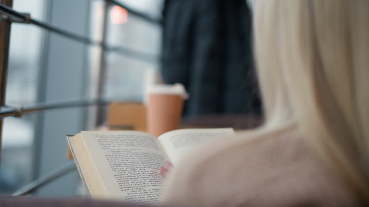 책에서 새로운 페이지로 전환하는 여자의 클로즈업 뒷면, 앞의 테이블에 차 컵, 몰에서 초점 밖의 사람들을 보여주는 흐릿한 배경