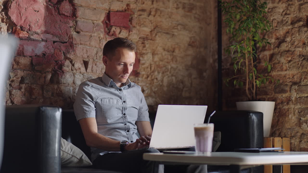 A young man in a shirt is sitting at a table with a laptop and typing on the keyboard. A student can study remotely. A businessman conducts his business remotely