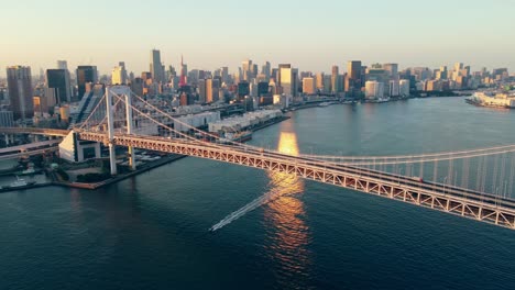 aerial drone flying low over water towards rainbow suspension bridge in odaiba tokyo city japan while boat sails under the bridge with tokyo tower in the background