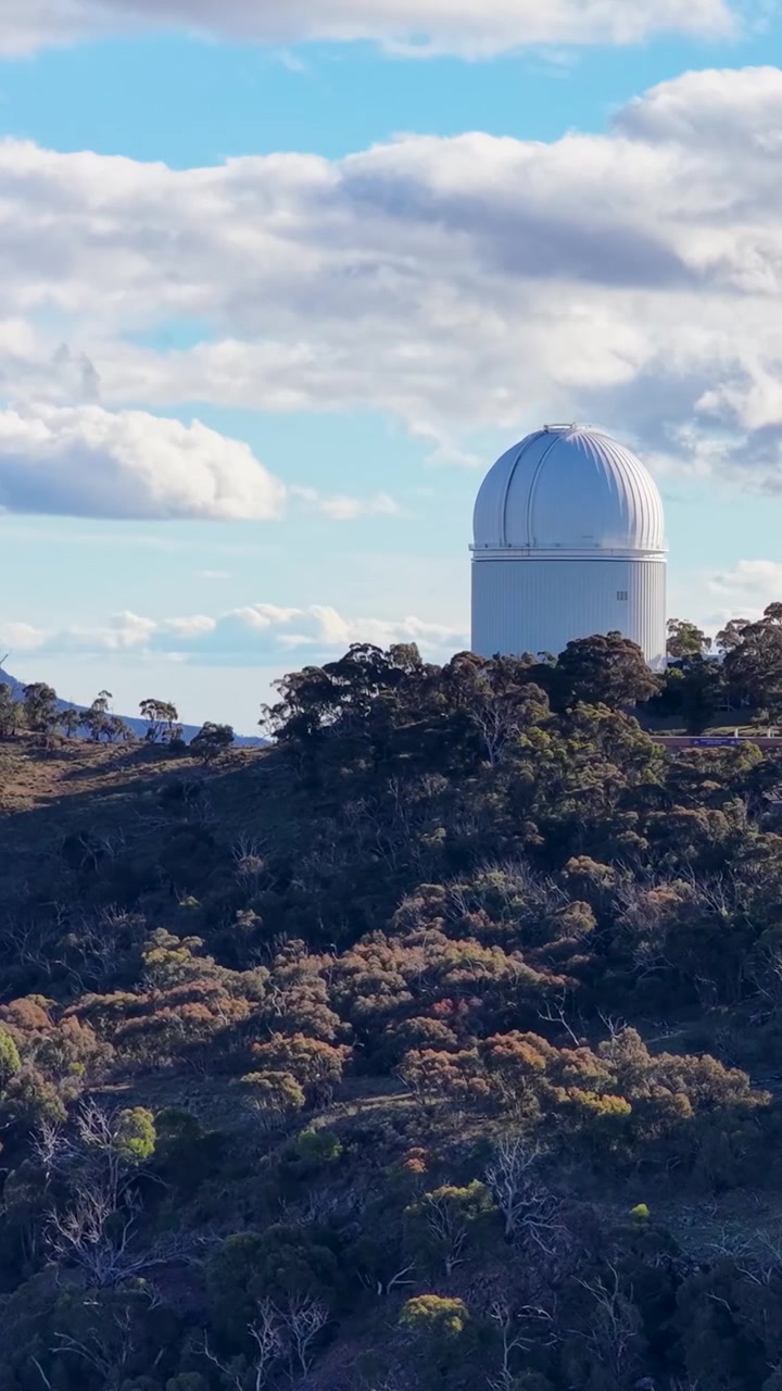 A large white astronomical observatory sits atop a tree-covered mountain ridge under cloudy skies.