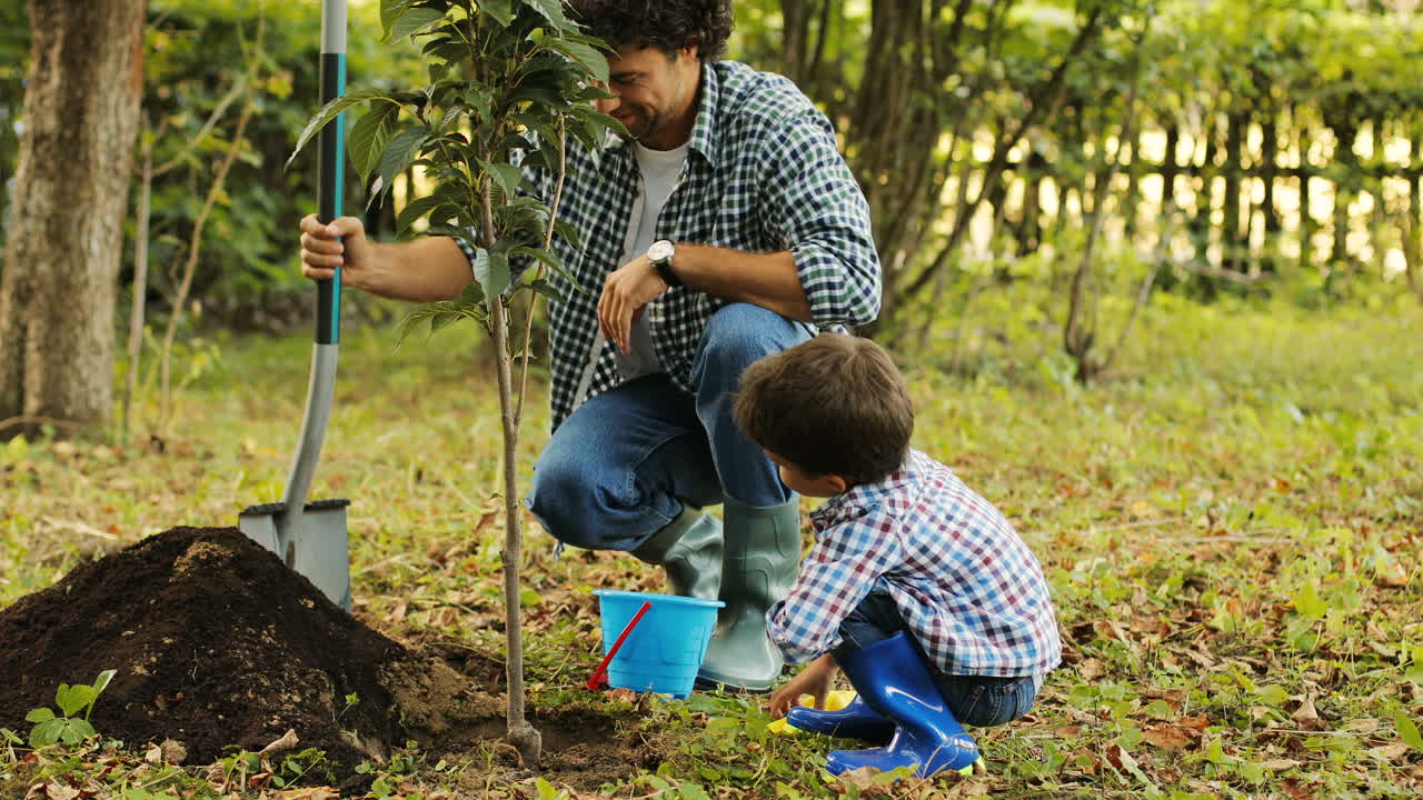 retrato de un niño y su padre plantando un árbol. el padre le sonríe a su hijo y cava la pala en el suelo. el niño juega con su cubo. fondo borroso