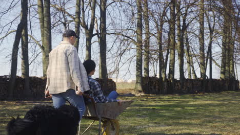 Caucasian-man-carrying-his-partner-on-a-wheelbarrow-in-the-countryside.-Their-dog-walks-around-them