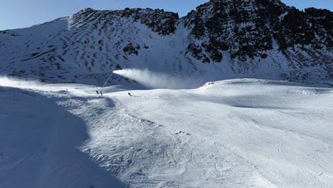 Multiple-skiers-coming-down-slope-with-active-snow-makers-and-rugged-cliffs-in-background