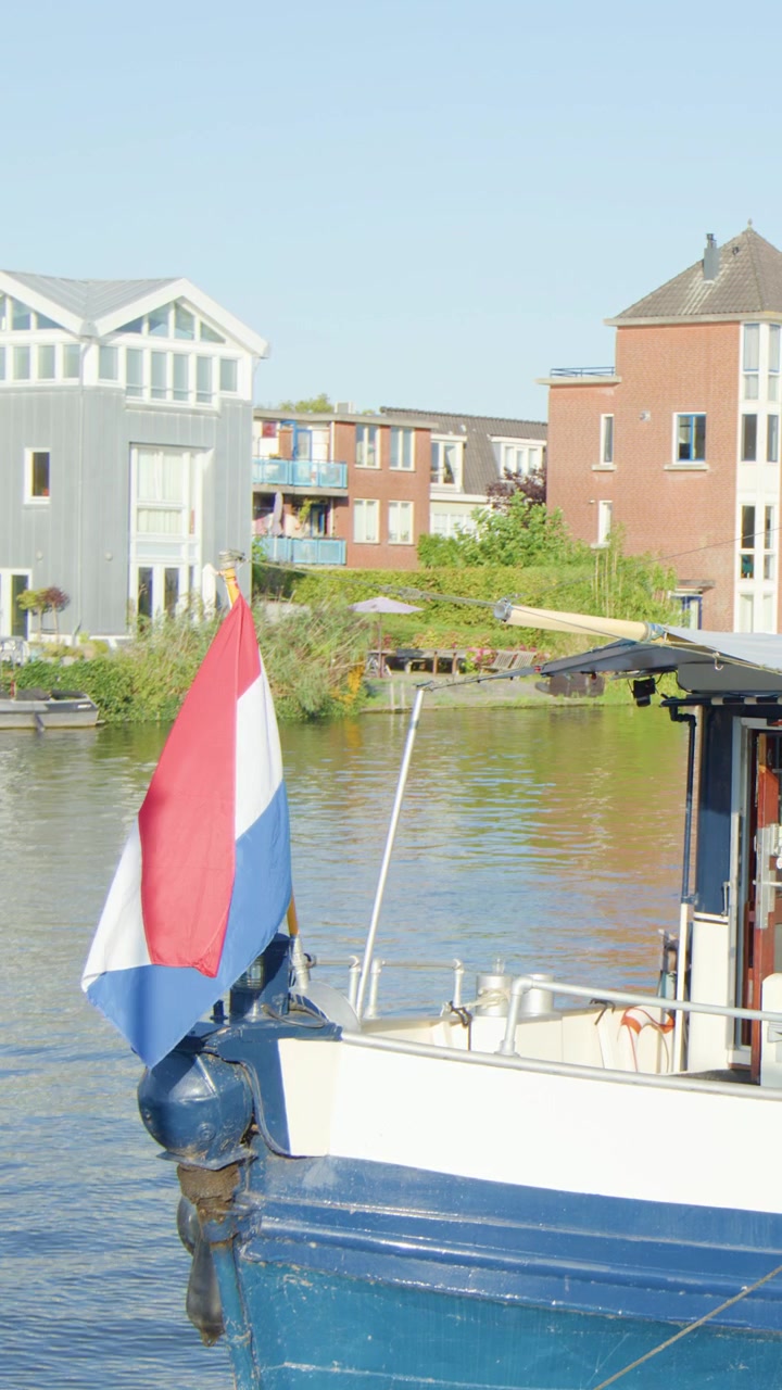 Bandiera olandese su una barca da canale blu e bianca ad Haarlem, Paesi Bassi, con moderne abitazioni sul lungocanale.