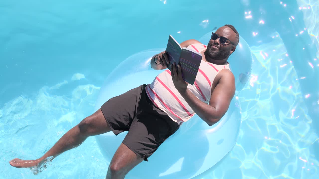 African American man reclining on blue float in pool reading book turning pages kicking relaxing