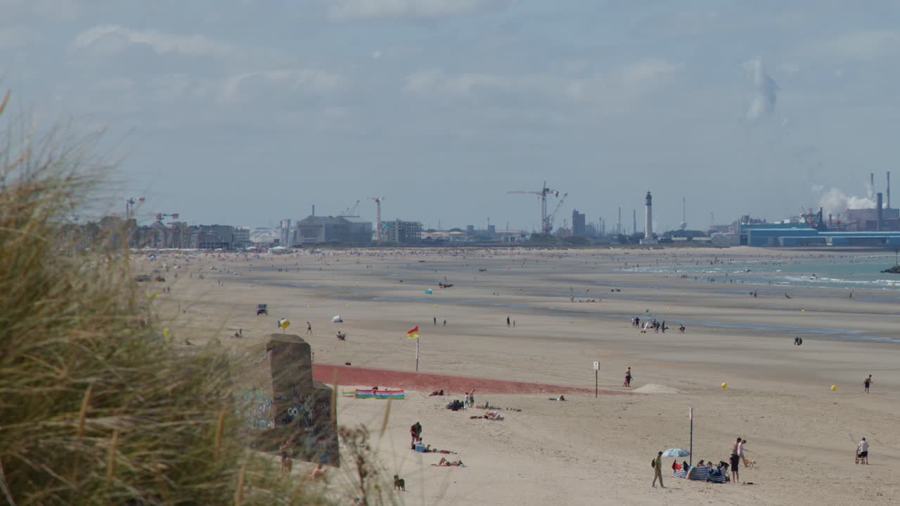 Wide shot of people on sandy beach with industrial port skyline in calm summer daylight