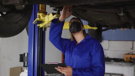 mecánico femenino con máscara facial tomando notas en el clipboard debajo de un coche en una estación de servicio de coches