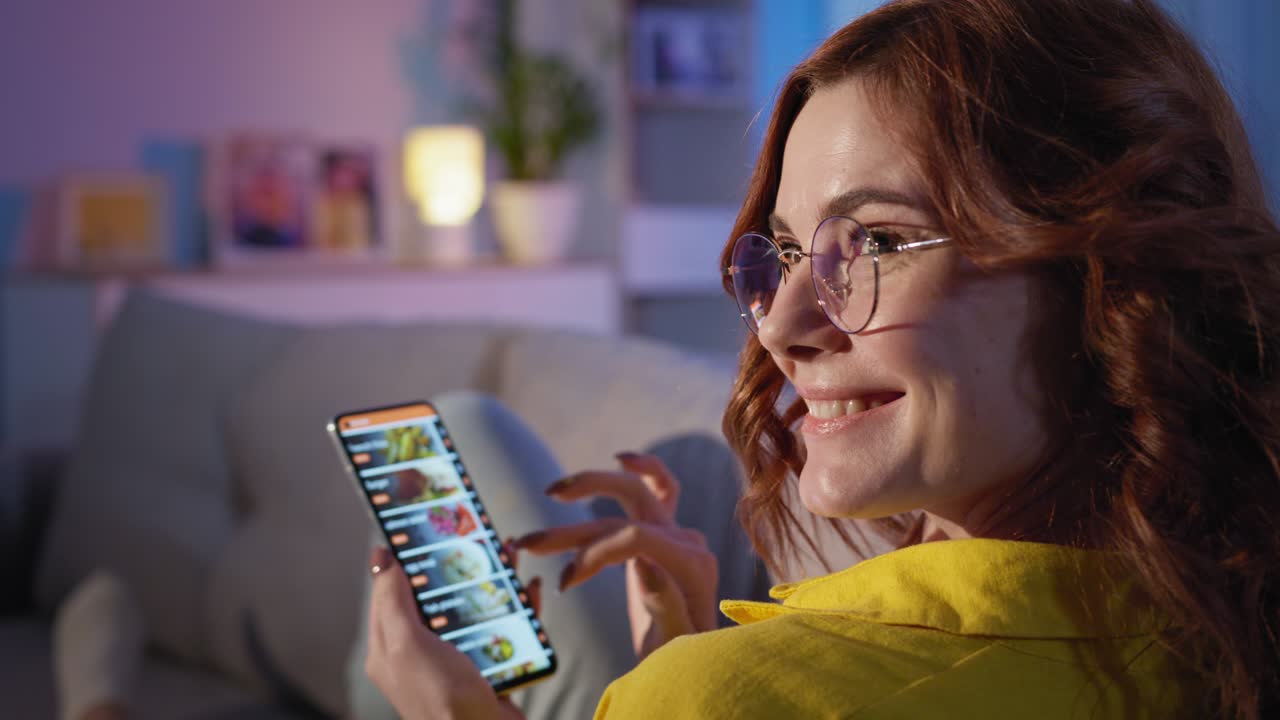 entrega a domicilio, joven cliente femenina sonriente con teléfono móvil en las manos ordena comestibles en una tienda en línea mientras se relaja en el sofá en la habitación por la noche