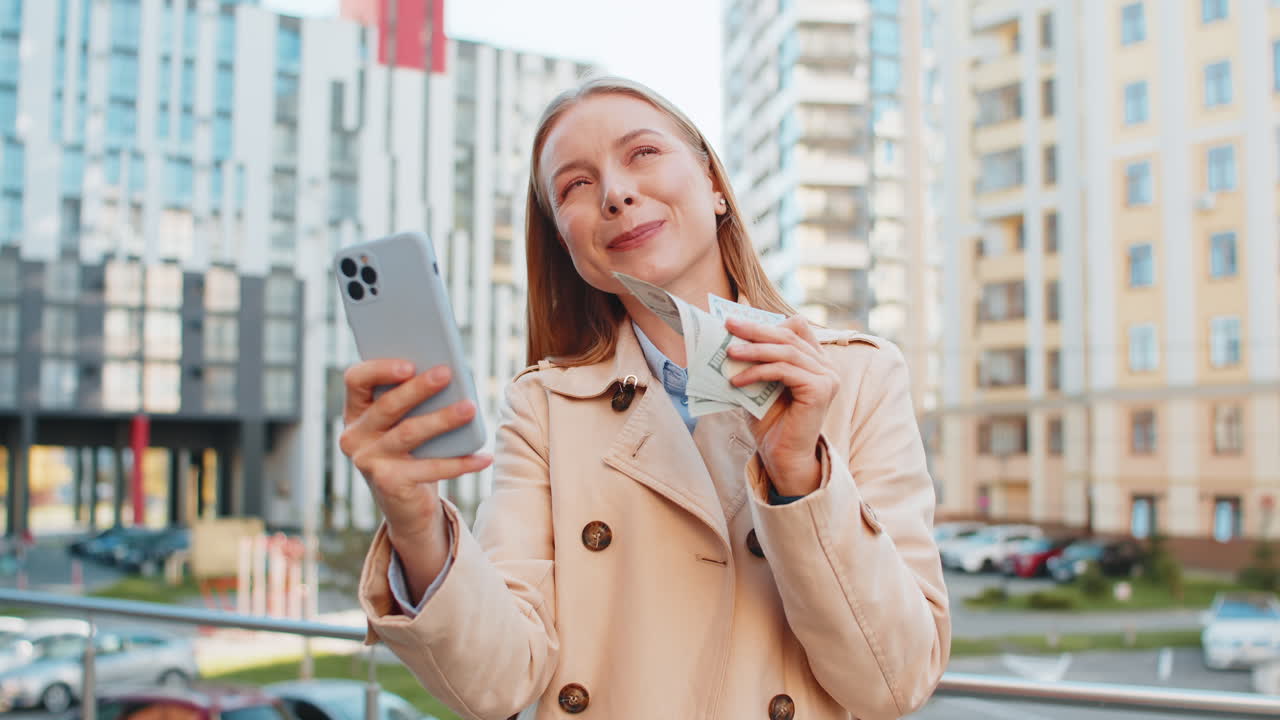 Happy rich mature caucasian businesswoman using smartphone and holding money on downtown city street
