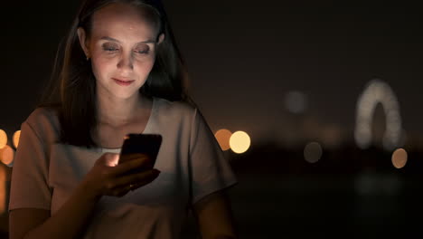 Una-Hermosa-Joven-En-Verano-En-Una-Gran-Ciudad-Nocturna-Mantiene-Un-Teléfono-Inteligente-En-Su-Interior-Y-Escribe-Un-Mensaje-Lee-Correos-Electrónicos-Y-Se-Comunica-En-Salas-De-Chat-Y-Redes-Sociales-Mantiene-Un-Blog-Mira-La-Pantalla-Del-Teléfono