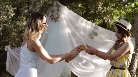 two ladies are resting in nature. fooling around. together they break the baguette. laughing, having fun. white cloth hanging on tree