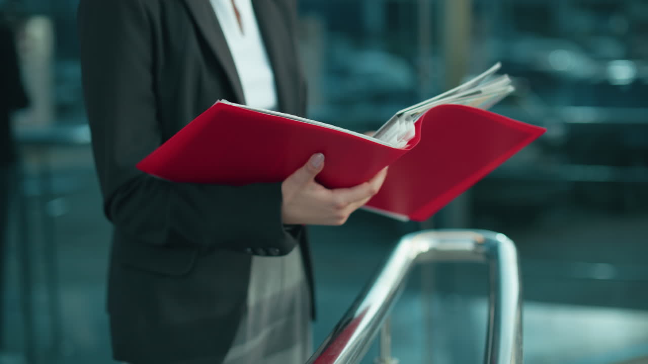 Partial view of woman in black blazer holding red folder while reviewing documents near modern urban setting with reflective residential building background and chrome railing under daylight