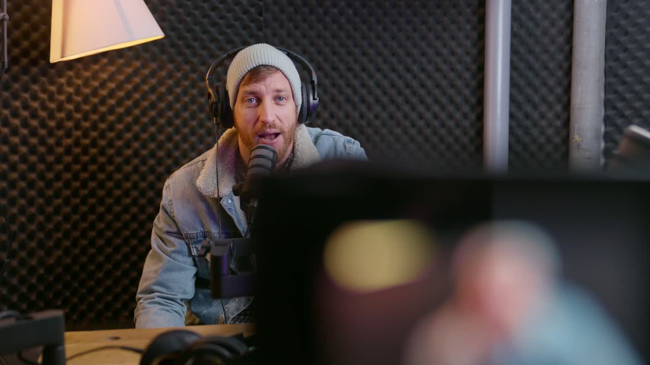 Professional podcast host or radio dj speaker talks to video camera while in sound studio. Using microphone and audio gear he films himself in sound proof booth. Excited and happy to stream live show