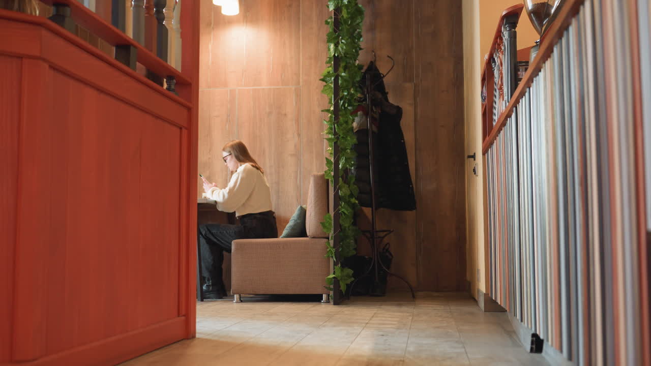 Woman seated alone in eatery operating phone with relaxed posture, surrounded by modern kitchen equipment and wall mounted cloth hanger in background