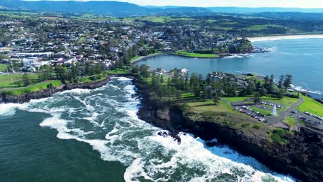 Drone-aerial-landscape-of-Kiama-harbour-lighthouse-and-blowhole-rural-town-suburbs-residential-housing-rocky-headland-waves-crashing-in-bay-South-Coast-travel-tourism