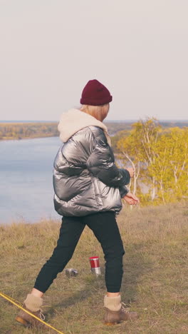 pretty girl tourist stretches body at tent and burning bonfire on river bank against clear sky in autumn morning