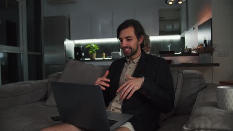 A-happy-brunette-man-with-stubble-in-a-black-jacket-and-beige-shorts-sits-on-the-sofa-and-communicates-via-video-conference-using-a-laptop-while-his-brunette-girlfriend-relaxes-in-the-kitchen-in-a-modern-studio-apartment