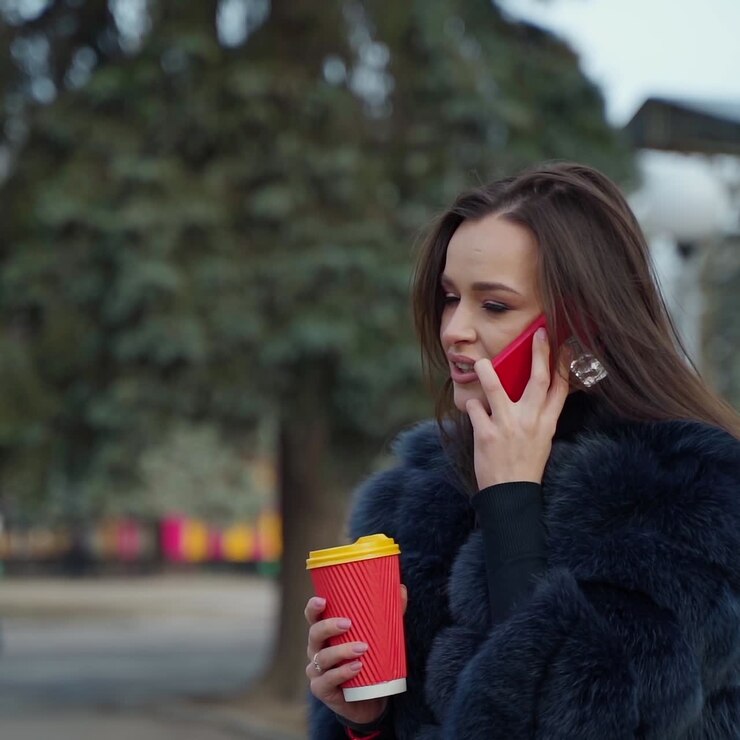 Charming girl in fluffy coat. Beautiful young brunette talk the mobile phone and smiles on blur park background. Woman in fur coat holds plastic cup of coffee outdoors.