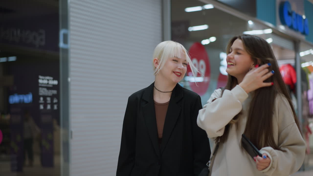 Two friends walk past clothing stores inside shopping mall, smiling and laughing while talking to each other, enjoying casual moment together under bright lights of modern retail environment