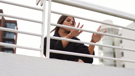 misteriosa mujer bonita con grandes ojos marrones con chaqueta negra y camiseta blanca se esconde detrás de la barandilla de la valla blanca tocándola luciendo linda impecable alrededor de hermosas casas en una ciudad a cámara lenta