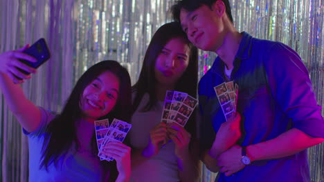 group of friends taking selfie having fun looking at prints from photo booth in nightclub or funfair 1