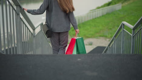 back view of lady in grey clothing holding colorful shopping bags while walking down stairs, hand gripping railing for support, with blurred greenery in the background