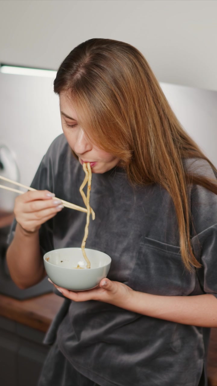 Primo piano di donna che mangia noodles con le bacchette mentre tiene una ciotola. La scena mostra dettagli della texture e illuminazione soffusa in un ambiente interno, concentrandosi sulle sue mani mentre la donna solleva il cibo dal bianco.
