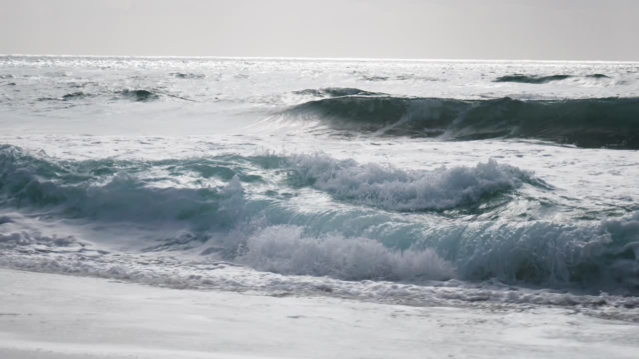 Rhythmic waves gently crash onto the shore, creating a soothing symphony of sound. The foamy surf dances across the golden sand, inviting beachgoers to enjoy the calming beauty of the ocean.