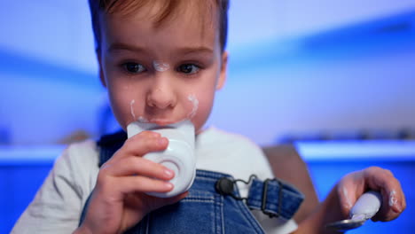 lovely kid holds an empty yogurt cup with his teeth. dairy spots are on boy's nose and cheeks. low angle view.
