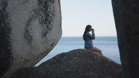 Junge-Attraktive-Touristin-Blickt-Auf-Das-Meer-Und-Fotografiert-Mit-Der-Kamera-Und-Genießt-Die-Friedliche-Aussicht-Auf-Das-Meer,-Während-Sie-Auf-Einem-Felsen-Sitzt-Und-Einen-Erholsamen-Urlaub-Genießt